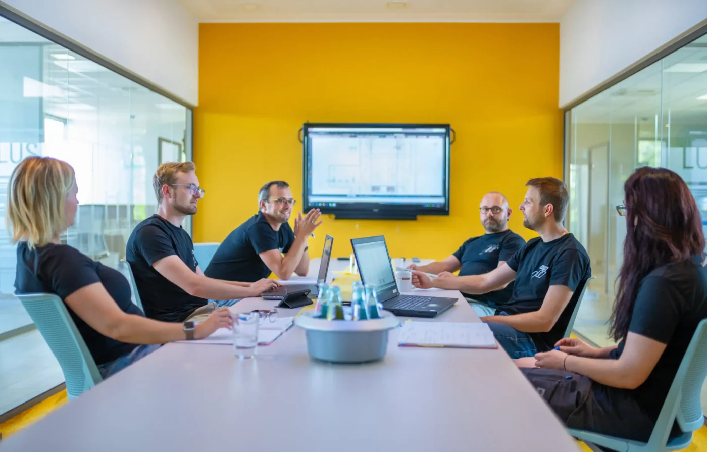 Mehrere Menschen sitzen an einem Tisch bei einem Meeting und unterhalten sich. Im Hintergrund ist ein Bildschirm zu sehen, auf dem Dateien angezeigt werden.